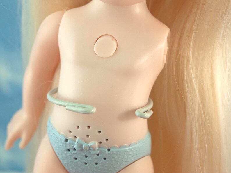 K&ouml;nnte beinhalten: Ein Puppenoberk&ouml;rper mit blonden Haaren und einem hellblauen Bikini-Unterteil. Die Puppe hat eine kreisf&ouml;rmige &Ouml;ffnung auf der Brust und ein hellblaues, gebogenes Accessoire um die Taille. Das Bikini-Unterteil hat eine Schleife.