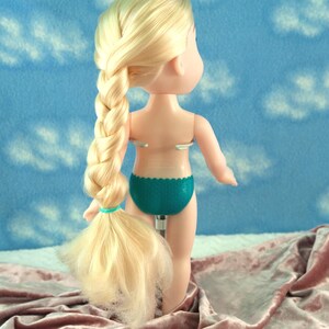 May include: A doll with blonde hair styled in a long braid. The doll is wearing teal underwear. The background is a blue fabric with white cloud shapes. The doll is standing on a pink fabric.
