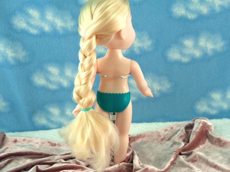 May include: A doll with blonde hair styled in a long braid. The doll is wearing teal underwear. The background is a blue fabric with white cloud shapes. The doll is standing on a pink fabric.