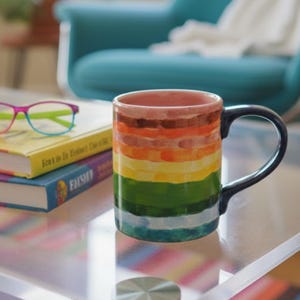 Caneca de cerâmica artesanal pintada à mão com listras do arco-íris e alça azul-marinho.
