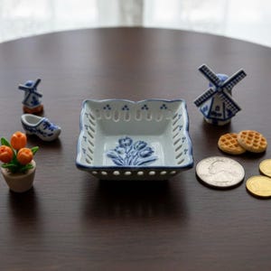 Może przedstawiać: Kolekcja miniaturowych przedmiotów w stylu holenderskim, w tym kwadratowa biało-niebieska miska z motywem tulipanów, wiatrak, chodak i doniczka z pomarańczowymi kwiatami. W zestawie również dwa gofry i kilka monet.