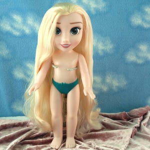 May include: A doll with long blonde hair and blue eyes, wearing a teal bikini bottom. The doll is standing on a pink velvet surface, with a blue background featuring white cloud shapes.