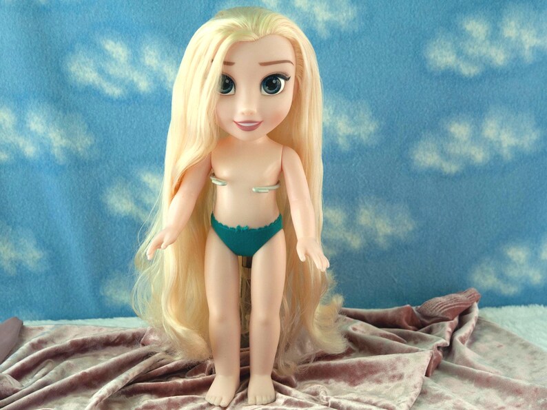 May include: A doll with long blonde hair and blue eyes, wearing a teal bikini bottom. The doll is standing on a pink velvet surface, with a blue background featuring white cloud shapes.