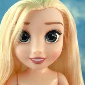 May include: A close-up of a doll's face with long, light blonde hair. The doll has large blue eyes with black eyelashes, a small nose, and a smiling mouth. The doll's skin is a pale peach color.