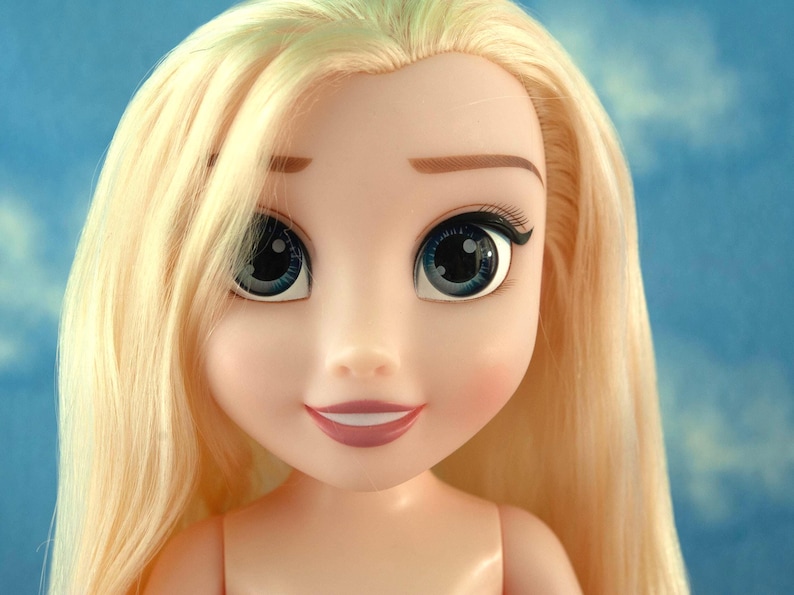 May include: A close-up of a doll's face with long, light blonde hair. The doll has large blue eyes with black eyelashes, a small nose, and a smiling mouth. The doll's skin is a pale peach color.