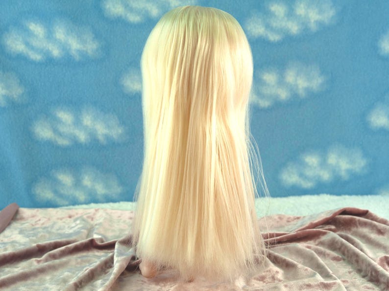 K&ouml;nnte beinhalten: Eine blonde Per&uuml;cke mit langem, glattem Haar. Die Per&uuml;cke wird vor einem blauen Hintergrund mit wei&szlig;en Wolkenmustern und einer rosa Samtoberfl&auml;che pr&auml;sentiert.