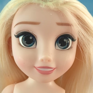 K&ouml;nnte beinhalten: Nahaufnahme eines Puppengesichts mit blonden Haaren und blauen Augen. Die Puppe hat einen hellen Teint mit gemalten Augenbrauen, Wimpern und einem dezenten rosa Rouge auf den Wangen. Die Lippen sind hellrosa bemalt, und die Puppe l&auml;chelt.