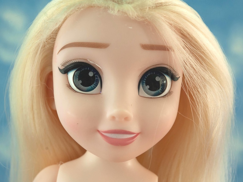 K&ouml;nnte beinhalten: Nahaufnahme eines Puppengesichts mit blonden Haaren und blauen Augen. Die Puppe hat einen hellen Teint mit gemalten Augenbrauen, Wimpern und einem dezenten rosa Rouge auf den Wangen. Die Lippen sind hellrosa bemalt, und die Puppe l&auml;chelt.
