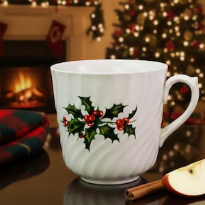 May include: A white ceramic mug with a holly and berry design. The mug has a curved handle and a swirled texture. A slice of apple and cinnamon sticks are on the table. The background includes a fireplace and Christmas tree.