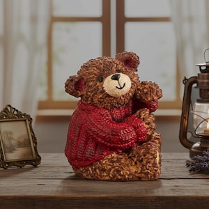 May include: A brown teddy bear figurine wearing a red sweater. The bear has a smiling face and is sitting on a wooden surface. A vintage-style lantern and framed picture are in the background.
