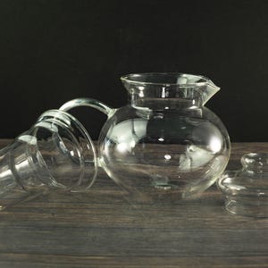 Puede incluir: Un juego de tetera de vidrio transparente con cuerpo redondo, boquilla y asa. El juego incluye una tapa y un infusor de vidrio separado. Las piezas están dispuestas sobre una superficie de madera oscura.