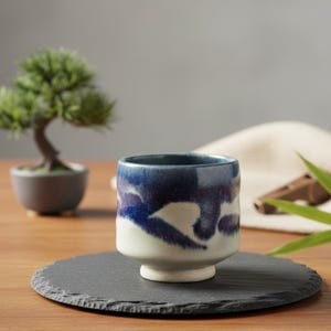 Puede incluir: Una taza de cerámica con un diseño abstracto azul y blanco. La taza está sobre un posavasos de pizarra negro. Un pequeño bonsái está en el fondo, junto con una tela blanca y un objeto de madera.