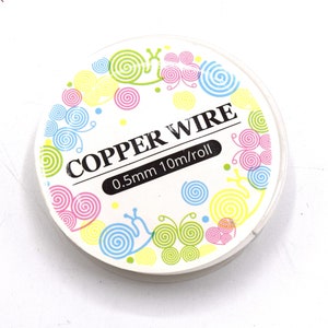 Pode incluir: Um rolo de fio de cobre com uma etiqueta branca. A etiqueta apresenta o texto "COPPER WIRE" e "0,5 mm 10 m/rolo". A etiqueta &eacute; decorada com desenhos coloridos de espirais e carac&oacute;is em rosa, azul, amarelo e verde.