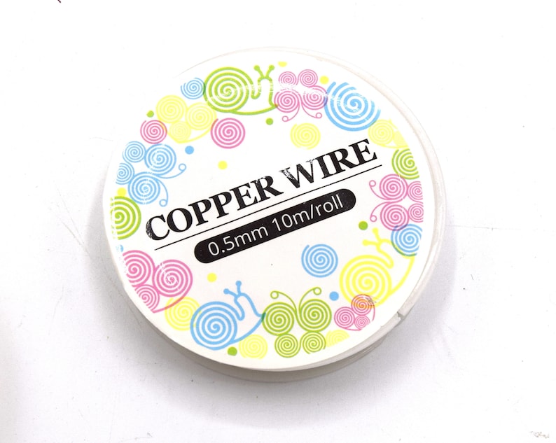 Pode incluir: Um rolo de fio de cobre com uma etiqueta branca. A etiqueta apresenta o texto "COPPER WIRE" e "0,5 mm 10 m/rolo". A etiqueta &eacute; decorada com desenhos coloridos de espirais e carac&oacute;is em rosa, azul, amarelo e verde.