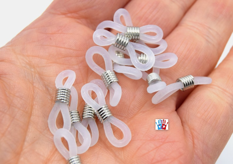 May include: A close-up of a pile of white plastic eyeglass end tips with silver metal rings. The end tips are designed to attach to the ends of eyeglass chains or cords.