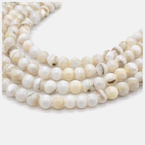 Peut inclure: Une collection de perles rondes et polies en nacre blanche, disposées en plusieurs rangées courbes. Chaque perle présente une surface lisse et irisée avec de subtiles variations de crème et de blanc cassé, reflétant la lumière avec un éclat nacré.