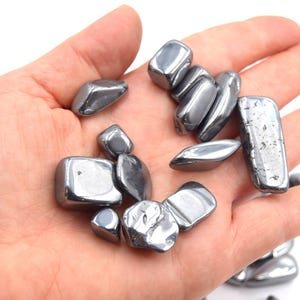 Peut inclure: Plusieurs pierres d'h&eacute;matite polies, de couleur gris m&eacute;tallique. Les pierres ont des formes vari&eacute;es, allant de rondes &agrave; rectangulaires, et refl&egrave;tent la lumi&egrave;re, cr&eacute;ant un aspect brillant. Les pierres sont lisses et semblent avoir &eacute;t&eacute; roul&eacute;es.