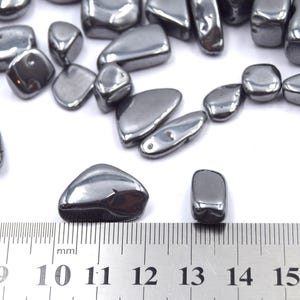 Peut inclure: Collection de pierres d'h&eacute;matite polies, gris fonc&eacute;, de formes et tailles vari&eacute;es. Les pierres ont un &eacute;clat m&eacute;tallique et sont dispos&eacute;es sur une surface blanche. Une r&egrave;gle avec des graduations en millim&egrave;tres est visible.