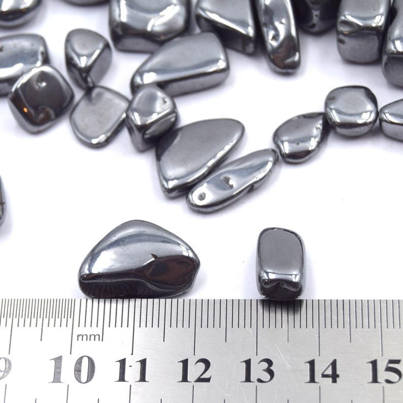 Peut inclure: Collection de pierres d'h&eacute;matite polies, gris fonc&eacute;, de formes et tailles vari&eacute;es. Les pierres ont un &eacute;clat m&eacute;tallique et sont dispos&eacute;es sur une surface blanche. Une r&egrave;gle avec des graduations en millim&egrave;tres est visible.