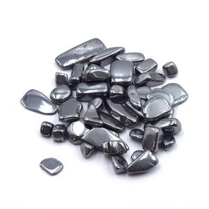 Peut inclure: Un tas de pierres d'h&eacute;matite polies, de couleur gris fonc&eacute;. Les pierres ont des formes vari&eacute;es, de rondes &agrave; rectangulaires, et une surface lisse et r&eacute;fl&eacute;chissante. Les pierres sont dispers&eacute;es sur un fond blanc.