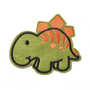 May include: A green felt dinosaur applique with orange embroidered details. The dinosaur has a spiky back and a cute, cartoon-like face.