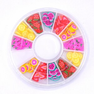 Peut inclure: Une roue en plastique blanche avec huit compartiments remplis de charmes pour ongles en forme de fruits colorés. Les charmes sont en forme de fraises, de tranches de pastèque, de citrons et de raisins.