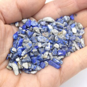 Może przedstawiać: Garść małych, niebieskich i białych odłamk&oacute;w lapis lazuli. Odłamki są nieregularne pod względem kształtu i rozmiaru, a niekt&oacute;re mają metaliczny połysk.