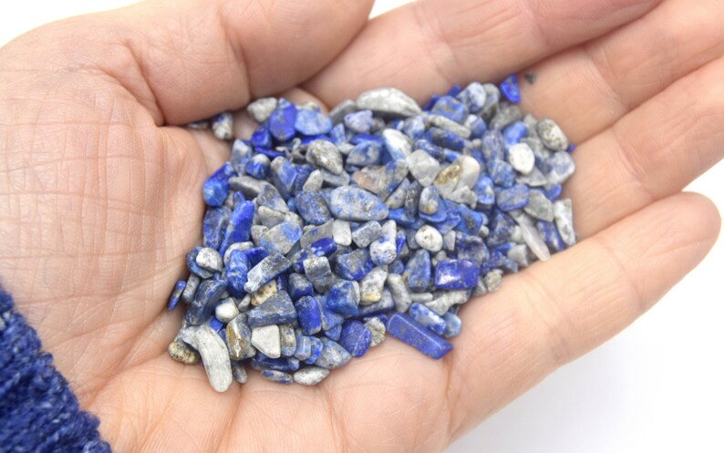 Może przedstawiać: Garść małych, niebieskich i białych odłamk&oacute;w lapis lazuli. Odłamki są nieregularne pod względem kształtu i rozmiaru, a niekt&oacute;re mają metaliczny połysk.