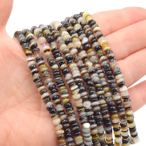 Pode incluir: Várias fileiras de pequenas contas redondas em tons de preto, cinza, branco e amarelo são seguradas em uma mão. As contas parecem ser feitas de um material natural e são enfiadas. As contas têm aproximadamente 0,6 cm de diâmetro.