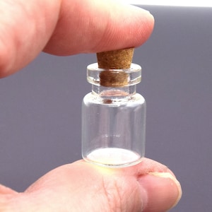 May include: A small, clear glass bottle with a cork stopper. The bottle is held between fingers, showcasing its compact size. The cork is a light brown color, contrasting with the transparent glass. Ideal for storing small items or as a decorative element.