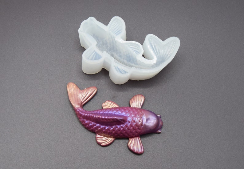 K&ouml;nnte beinhalten: Eine Silikonform zum Herstellen eines violetten Koi-Fischs. Die Form ist ge&ouml;ffnet und ein fertiger Koi-Fisch aus Harz ist daneben abgebildet.