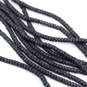 Peut inclure: Gros plan sur plusieurs brins de perles de roche volcanique noires. Les perles sont cylindriques avec une surface texturée et sont enfilées ensemble. Le fond est blanc, mettant en valeur la couleur foncée des perles. Idéal pour la création de bijoux.