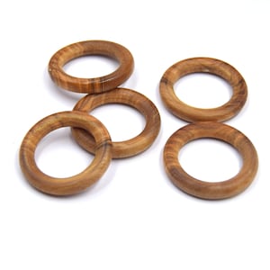 Könnte beinhalten: Fünf Holzringe, jeder mit einem anderen Maserungsmuster, liegen auf einer weißen Oberfläche. Die Ringe sind alle gleich groß und gleich geformt und aus hellem braunem Holz gefertigt.