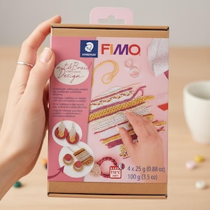 Op de afbeelding: Een knutseldoos met de tekst "Knit & Braid Design" en de merknaam "FIMO". De doos is roze en bruin, met afbeeldingen van afgewerkte brei- en vlechtprojecten. De set bevat 4 x 25 g boetseerklei.