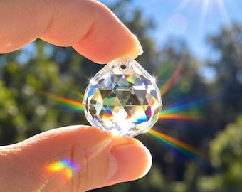 Könnte beinhalten: Ein klarer, facettierter Glaskristallanhänger mit einem kleinen Loch an der Oberseite zum Aufhängen. Der Kristall bricht das helle Sonnenlicht und erzeugt lebendige Regenbogenlichtreflexe und Sternmuster, die von seiner Oberfläche ausgehen.