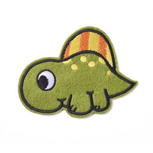 Op de afbeelding: Groene vilt dinosaurus applicatie met een gestreepte oranje en gele kam. De dinosaurus heeft een groot oog met een zwarte pupil en een witte iris.