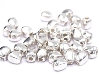 Pode incluir: Um monte de contas de metal prateado com forma de lágrima e um furo no centro. As contas estão espalhadas sobre uma superfície branca.