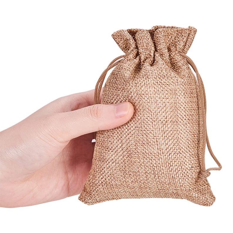 May include: A small, brown burlap drawstring bag with a natural, woven texture. The bag is held in a hand, showcasing its size and shape.
