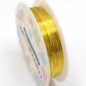 Pode incluir: Uma bobina de fio de cobre dourado, com 0,5 mm de espessura, &eacute; exibida sobre um fundo branco. A lateral da bobina apresenta desenhos em espiral coloridos e o texto "COPPER WIRE". Este fio &eacute; adequado para projetos de joalheria e artesanato.