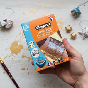 Könnte beinhalten: Eine Schachtel Cléopâtre Glass Flex Epoxidharz mit Text auf Französisch, Deutsch und Spanisch. Die Schachtel ist orange und blau, gehalten von einer Hand. Die Schachtel zeigt Beispiele für die Verwendung des Harzes. Das Produkt enthält 130 ml.