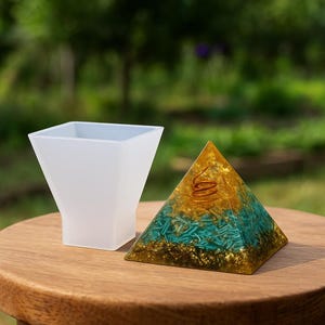 Può includere: Un contenitore bianco, quadrato e traslucido accanto a una piramide decorativa. La piramide presenta accenti dorati, turchesi e ramati. La piramide è realizzata in resina e ha una spirale di rame sulla parte superiore. Gli oggetti sono su una superficie di legno.