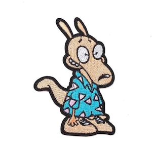 Può includere: Toppa ricamata raffigurante un personaggio dei cartoni animati con corpo marrone chiaro, occhi grandi e camicia blu con motivi triangolari rosa. La toppa ha un bordo nero e si trova su sfondo bianco.