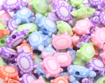 May include: A close-up of a pile of colorful plastic turtle beads. The beads are in various shades of pink, purple, blue, green, and orange. Each bead has a white outline of a turtle shell.