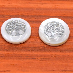 Puede incluir: Dos botones redondos blancos con un diseño de árbol de la vida plateado. Los botones están sobre una superficie de madera.