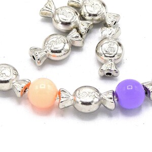 Peut inclure: Un bracelet à breloques argenté avec des breloques en forme de bonbons qui disent "Love" et "Heart". Le bracelet comprend également deux perles rondes, une orange et une violette.