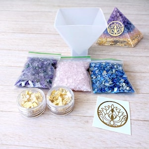 Può includere: Un kit artigianale con un imbuto bianco, sacchetti di pietre viola, rosa e blu, due vasetti di scaglie d'oro, una piramide e un adesivo con un disegno di loto dorato. La piramide ha un disegno di loto dorato ed è viola, blu e oro.