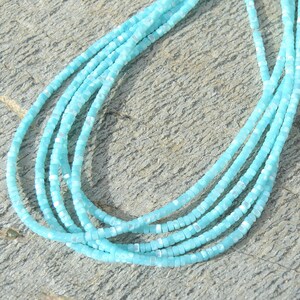 Pode incluir: Um fio de contas quadradas azuis claras com um leve brilho iridescente. As contas são enfiadas em um fio transparente e estão dispostas em um padrão circular.