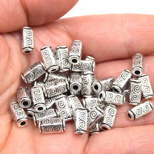 Peut inclure: Un tas de perles en métal argenté avec un motif en spirale. Les perles sont de forme rectangulaire et ont un trou au centre pour le perlage.