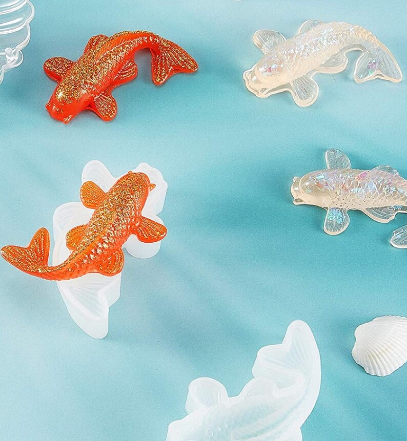 K&ouml;nnte beinhalten: F&uuml;nf Harz-Goldfisch-Ornamente in verschiedenen Posen. Zwei sind orange mit goldenem Glitzer, zwei sind klar mit schimmerndem Glitzer und eines ist klar mit einer wei&szlig;en Muschel.