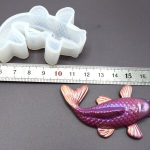 K&ouml;nnte beinhalten: Eine transparente Silikonform eines Koi-Fischs, etwa 10 cm lang, neben einem in Lila und Gold bemalten Koi-Fisch aus Harz.